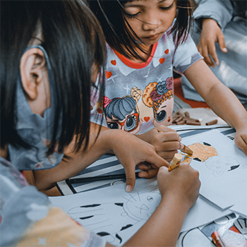 Two children sitting at a desk coloring images of a cat